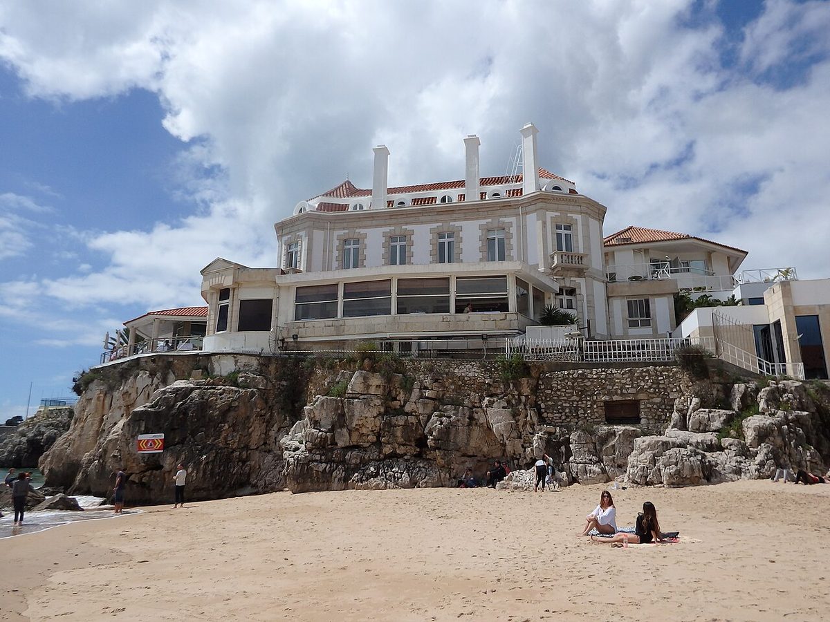 Sunbathers on the sand at Praia da Conceição in Cascais below a white clifftop villa