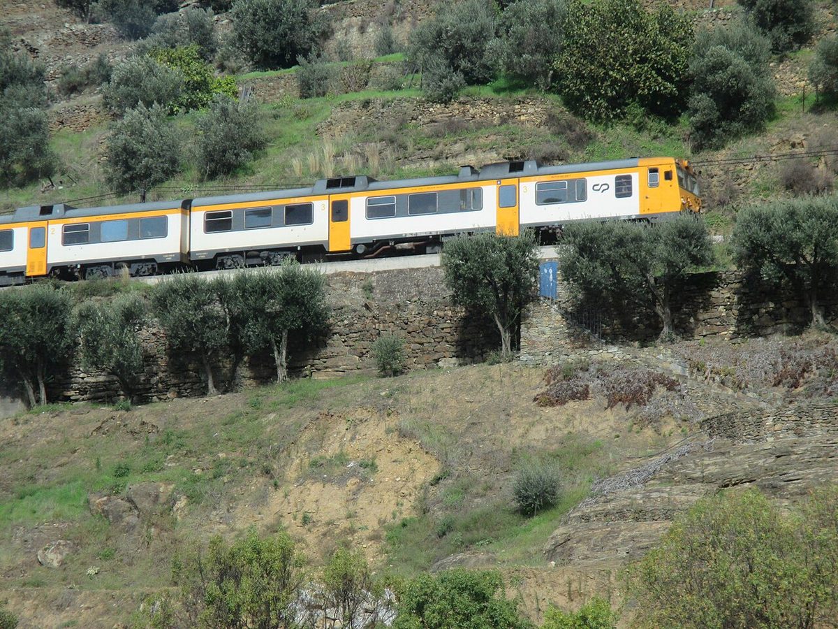 A yellow and white CP regional train running along the Linha do Douro above terraced hillsides